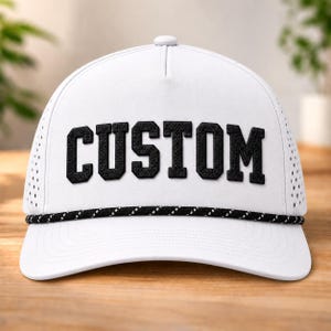 May include: A white baseball cap featuring the word CUSTOM in bold black embroidered lettering across the front panels. The cap includes a black and white braided rope detail along the base of the crown and perforated side panels for ventilation.