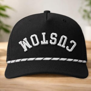 K&ouml;nnte beinhalten: Schwarze Baseballkappe mit dem Wort "CUSTOM" in wei&szlig;en Blockbuchstaben gestickt. Die Kappe hat ein wei&szlig;es und schwarzes Seildetail am Rand und kleine Bel&uuml;ftungsl&ouml;cher an den Seiten.