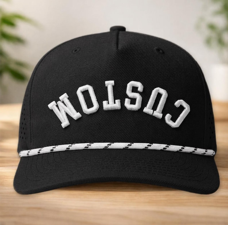 K&ouml;nnte beinhalten: Schwarze Baseballkappe mit dem Wort "CUSTOM" in wei&szlig;en Blockbuchstaben gestickt. Die Kappe hat ein wei&szlig;es und schwarzes Seildetail am Rand und kleine Bel&uuml;ftungsl&ouml;cher an den Seiten.