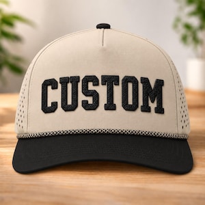 Puede incluir: Una gorra de béisbol beige con visera negra y un detalle de cuerda negra en la base de la corona. El frente presenta la palabra CUSTOM en letras de bloque negras, texturizadas y en negrita. Los paneles laterales tienen un patrón perforado.