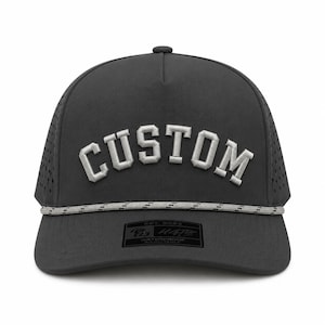 Puede incluir: Gorra impermeable gris oscuro con la palabra "CUSTOM" bordada en blanco en la parte delantera. La gorra tiene un detalle de cuerda y un peque&ntilde;o parche rectangular con texto. La gorra tiene una visera curva y un dise&ntilde;o de malla en los laterales.