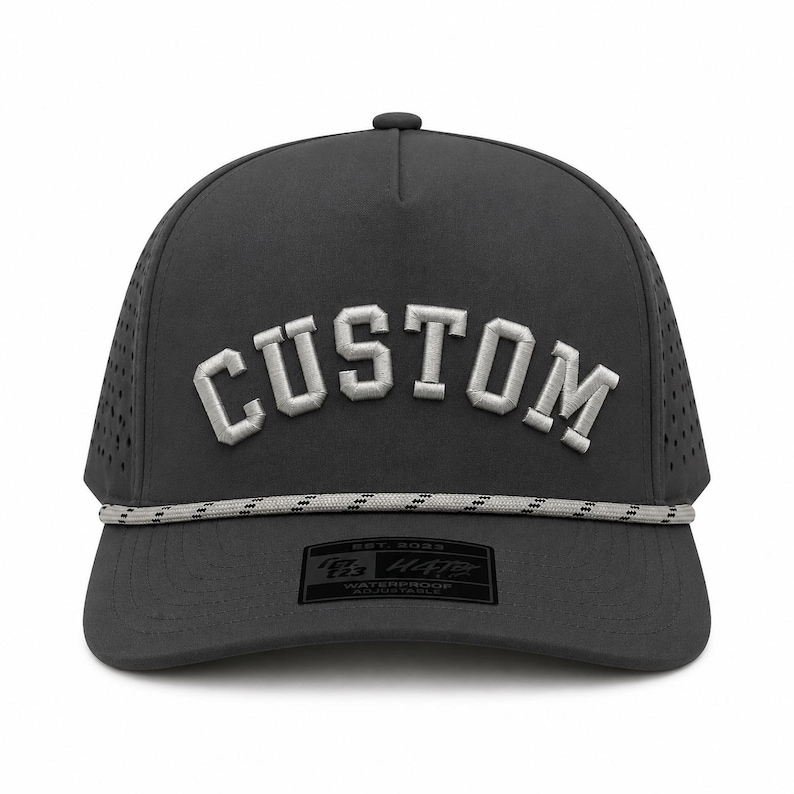 Puede incluir: Gorra impermeable gris oscuro con la palabra "CUSTOM" bordada en blanco en la parte delantera. La gorra tiene un detalle de cuerda y un peque&ntilde;o parche rectangular con texto. La gorra tiene una visera curva y un dise&ntilde;o de malla en los laterales.
