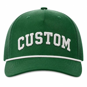 Puede incluir: Una gorra tipo camionero de color verde bosque con un detalle de cuerda blanca en la base de la corona y la palabra CUSTOM bordada en letras blancas de bloque en el panel frontal. La gorra tiene un frente estructurado y malla trasera.