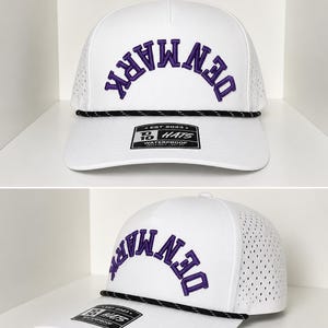 K&ouml;nnte beinhalten: Wei&szlig;e Baseballkappe mit dem Wort "DENMARK" in Lila auf der Vorderseite gestickt. Die Kappe hat ein schwarz-wei&szlig;es Seildetail und einen gebogenen Schirm. Die Kappe hat ein perforiertes Seitenteil und ein Etikett mit der Aufschrift "WATERPROOF".