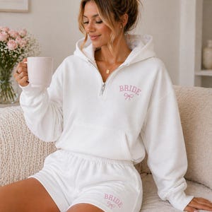 Könnte beinhalten: Ein weißes Braut-Set, bestehend aus einem Hoodie mit Reißverschluss und passenden Shorts. Das Wort "BRIDE" ist in rosa Schrift mit einer Schleife auf dem Hoodie und den Shorts aufgedruckt. Das Outfit ist aus weichem Material.