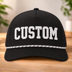 以下が含まれることがあります： 前面に大きな白いブロック体で「CUSTOM」という文字が刺繍された黒い野球帽。帽子のクラウンの付け根には白いロープのディテールがあり、通気のためのパンチング加工が施されたサイドパネルと、カーブしたつばが特徴です。