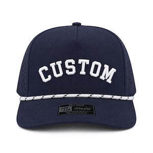 Puede incluir: Gorra de b&eacute;isbol azul marino con la palabra "CUSTOM" en blanco, arqueada en la parte delantera. La gorra tiene un detalle de cuerda blanca y negra a lo largo del borde y una peque&ntilde;a etiqueta que dice "10/10 Hats".