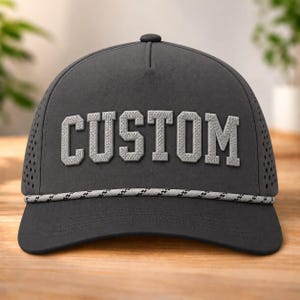 以下が含まれることがあります： 前面に盛り上がったライトグレーのブロック体で「CUSTOM」という文字がデザインされたダークグレーの野球帽。帽子のクラウンの付け根にはライトグレーと黒のツイストロープの装飾があり、サイドパネルには通気孔が施されています。