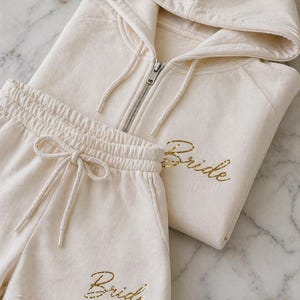 Puede incluir: Conjunto de ropa de estar por casa color crema con una sudadera con capucha y cremallera y pantalones cortos a juego. La palabra "Bride" está impresa en letras doradas en ambas piezas. Los pantalones cortos tienen cintura con cordón.