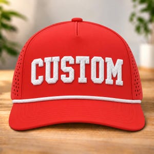 以下が含まれることがあります： フロントパネルに大きな白いテクスチャー加工のブロック体で「CUSTOM」という文字がデザインされた、鮮やかな赤色の野球帽。帽子のクラウンの付け根には白いロープのディテールがあり、側面には通気用のパンチング加工が施されています。