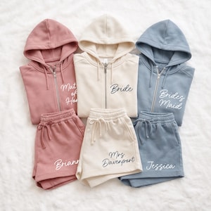 Puede incluir: Conjuntos a juego de sudaderas con capucha y cremallera y pantalones cortos en rosa empolvado, crema y azul. Las sudaderas llevan las palabras "Bride", "Bridesmaid" y "Matron of Honor". Los pantalones cortos están personalizados.