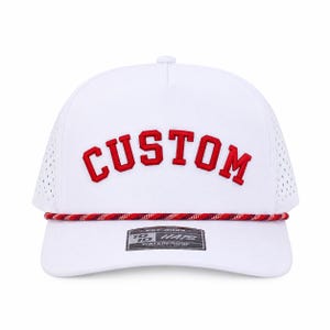 Puede incluir: Gorra de b&eacute;isbol blanca con la palabra "CUSTOM" bordada en rojo. La gorra tiene un detalle de cuerda roja y blanca y una peque&ntilde;a etiqueta que dice "10 HATS WATERPROOF".
