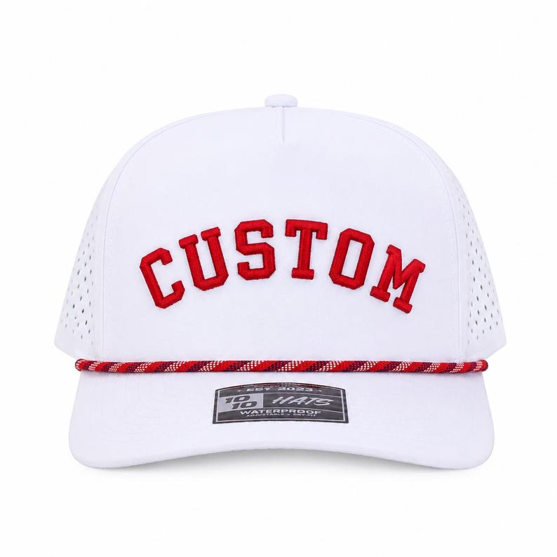 Puede incluir: Gorra de b&eacute;isbol blanca con la palabra "CUSTOM" bordada en rojo. La gorra tiene un detalle de cuerda roja y blanca y una peque&ntilde;a etiqueta que dice "10 HATS WATERPROOF".