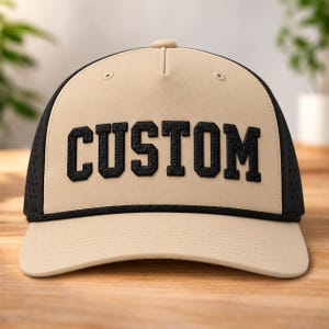 Puede incluir: Una gorra de béisbol beige con parte trasera de malla negra y un detalle de cuerda negra sobre la visera. El panel frontal presenta la palabra CUSTOM en letras de bloque negras, texturizadas y en negrita. La gorra tiene una corona estructurada.