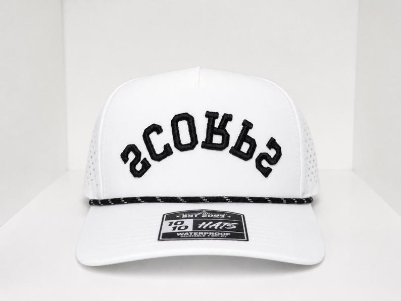 K&ouml;nnte beinhalten: Wei&szlig;e Baseballkappe mit dem Wort "SCOBBS" in Schwarz auf der Vorderseite gestickt. Die Kappe hat ein schwarz-wei&szlig;es Seildetail am Rand und ein kleines Etikett auf der Vorderseite. Der Hut ist wasserdicht.