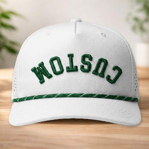 Può includere: Cappellino da baseball bianco con la scritta "CUSTOM" ricamata in lettere verdi. Il cappellino ha un dettaglio a corda verde e bianca lungo il bordo e piccoli fori di ventilazione sul lato.
