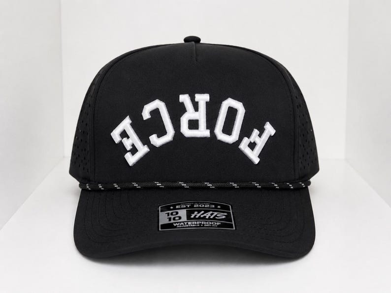 K&ouml;nnte beinhalten: Schwarze Baseballkappe mit dem wei&szlig;en Wort "FORCE" in einem Bogen &uuml;ber der Vorderseite. Die Kappe hat einen gebogenen Schirm, ein schwarz-wei&szlig;es Seildetail und ein kleines Etikett auf der Vorderseite. Die Kappe ist wasserdicht.