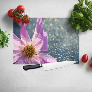 May include: A pink flower with a yellow center sits on a blue and white cutting board with a silver knife. There are red tomatoes and green basil leaves on the counter.