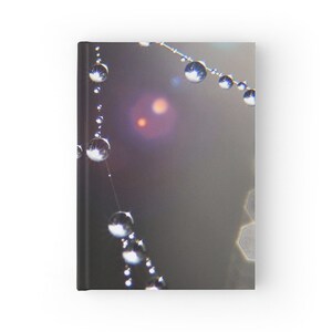 May include: Hardcover journal with a close-up of a spider web adorned with water droplets. The droplets reflect light, creating a sparkling effect against a blurred, dark background. The journal's cover is visible on the left side.