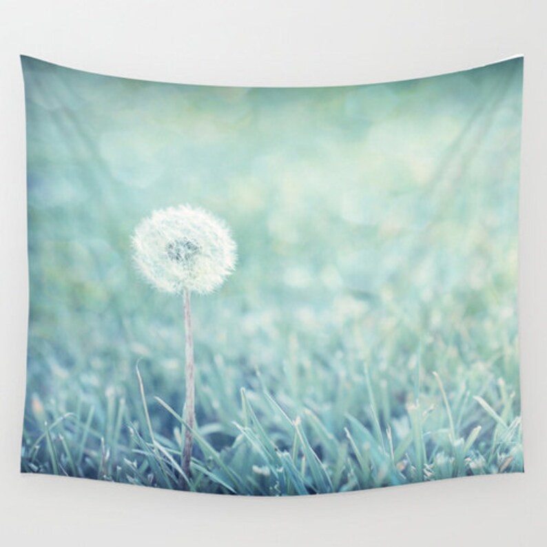 May include: A single dandelion seed head with a white, fluffy bloom stands tall in a field of soft, green grass. The background is a blurred, light blue and green color.