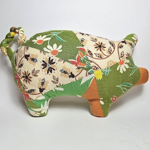 May include: A handmade patchwork pig-shaped pillow. The pig is made of various fabric scraps in green, cream, and orange, with floral and geometric patterns. The tail is a twisted rope of fabric. A unique decorative item.