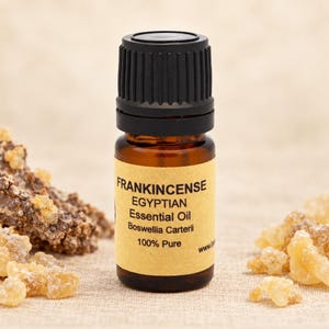 Op de afbeelding: Een klein amberkleurig glazen flesje met Egyptische wierook etherische olie en een zwarte geribbelde dop. Het beige etiket vermeldt FRANKINCENSE EGYPTIAN Essential Oil Boswellia Carterii 100% Pure. Het flesje is omringd door kleine stukjes hars.