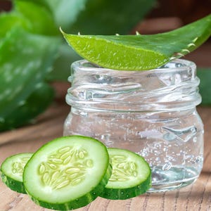 May include: A clear glass jar filled with a liquid, topped with a green aloe vera leaf. Sliced cucumbers are in front of the jar. The background features green leaves and a wooden surface.