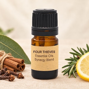 Op de afbeelding: Een klein amberkleurig glazen flesje met een zwarte geribbelde dop en een beige etiket met de tekst FOUR THIEVES Essential Oils Synergy Blend. Het flesje is omringd door kaneelstokjes, kruidnagels, een groen blad en een schijfje citroen.