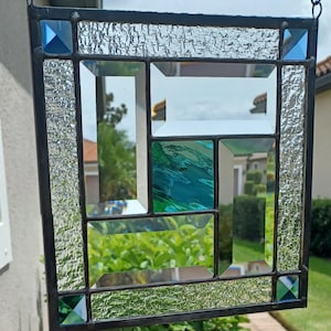 May include: Stained glass window panel with a geometric design. The panel features clear, textured, and colored glass pieces in shades of blue and green, framed by a dark metal border. The design includes squares and rectangles.
