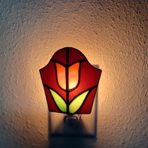May include: A stained glass night light featuring a red tulip design with yellow leaves, illuminated from within. The piece is constructed with dark metal soldering lines and is plugged directly into a wall outlet, casting a warm glow.