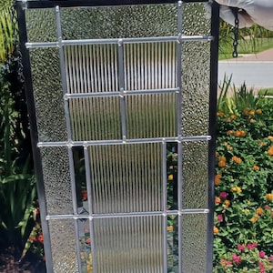 May include: A rectangular stained glass window with a black metal frame and a chain. The window features textured and ribbed glass panels. The window is held up by a gloved hand against a background of green foliage and flowers.