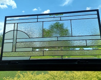 May include: A rectangular stained glass window with a black metal frame. The glass is clear with textured and smooth sections, and a semi-circle design. The window is positioned in front of a bright blue sky with clouds.