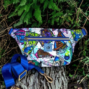 May include: A white fanny pack with a colorful print featuring Harry Potter themed images. The pack has a blue zipper and a blue adjustable strap. The strap has gold clasps. The pack is sitting on a tree trunk with green foliage in the background. The text "Creative Sister Designs Handmade" is visible in the lower right corner of the image.