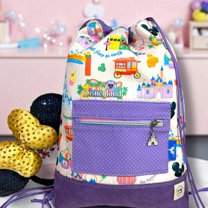 May include: A drawstring backpack with a Disneyland theme. The bag features a white background with colorful illustrations of park attractions, a purple polka-dot pocket, and a purple base. A silver castle charm hangs from the zipper.