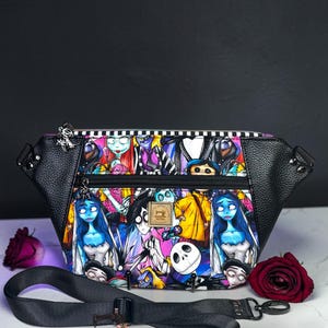 May include: A black faux leather fanny pack featuring a colorful printed fabric panel with various animated characters, including a blue-skinned bride and a white skull. The bag has a black zipper, a black adjustable strap, and a small square brand label.