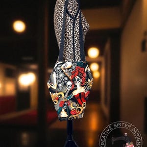 May include: A black tote bag with a Day of the Dead design featuring skulls, roses, and a woman in a red dress. The bag has black straps and is displayed on a mannequin with a leopard print pattern. The bag is handmade.