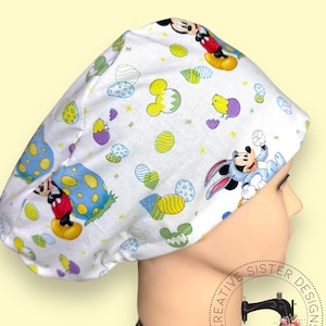 May include: White scrub hat with a Mickey Mouse Easter egg print. The hat features Mickey Mouse and a bunny rabbit with Easter eggs and chicks. The hat is made by Creative Sister Designs.