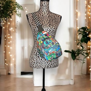 May include: A colorful, patterned crossbody bag with a black and white checkered strap. The bag is displayed on a mannequin with a leopard print torso.