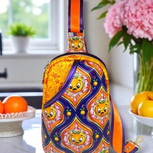 May include: A handmade sling bag with a vibrant orange, purple, and yellow pattern featuring a cartoon orange character. The bag has an orange and purple strap and a gold zipper. The text "creative sister designs handmade" is visible.