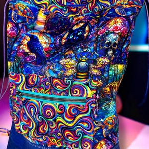 May include: A handmade backpack with a vibrant, psychedelic design. The bag features a blue base, a teal zipper pocket, and a drawstring closure. The colorful pattern includes swirls, a raven, a bee, a skull, and turtles. The text "creative sister designs handmade" is visible.