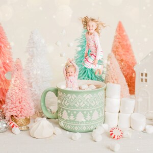 May include: Two children are jumping into a green mug filled with hot chocolate and marshmallows. The mug has a white snowflake pattern and is surrounded by miniature pink and white Christmas trees, marshmallows, and candy canes.