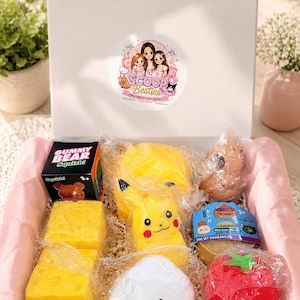 May include: A white gift box containing various squishy toys, including a yellow Pikachu figure, a red tomato, a white rice ball with a face, two yellow cheese blocks, a Gummy Bear box, and a Bun mystery set, all nestled in shredded paper.