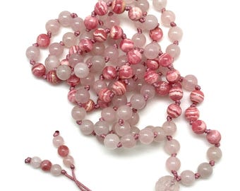 May include: A pink and white gemstone mala necklace with a knotted cord and a carved gemstone bead at the end.