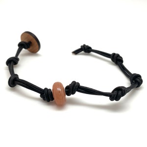 May include: A black leather bracelet with a wooden button clasp and a peach-colored bead. The bracelet is knotted between the bead and the clasp. The bracelet is on a white background.