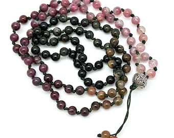 May include: A multi-coloured gemstone mala necklace with black, brown, pink, and green beads. The necklace is made with a dark green cord and has a silver charm at the end.