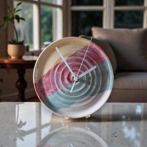 May include: A decorative ceramic clock with a unique design. The clock face is a plate with a spiral pattern in shades of pink, blue, and beige. The clock hands are white. The clock is displayed on a table.