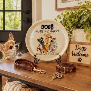May include: A cream-colored ceramic plate on a black metal stand features a cartoon illustration of various dogs and the text DOGS MAKE ME HAPPY HUMANS MAKE MY HEAD HURT. Nearby are a brown leather leash, a collar, and a wooden sign reading Dogs Welcome.