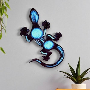 May include: A wall-mounted metal lizard sculpture in shades of blue and black. The lizard features a stylized design with glowing blue accents on its body and tail. The sculpture is mounted on a gray wall, with a plant in the background.