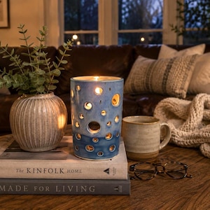 May include: A cylindrical blue ceramic candle holder with circular cutouts, a ribbed beige vase with green foliage, a speckled ceramic mug, and black-rimmed glasses resting on two stacked books titled The Kinfolk Home and Made for Living.