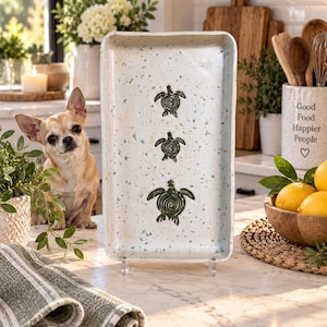 May include: A rectangular ceramic serving tray with a speckled off-white glaze featuring three dark green sea turtle illustrations arranged vertically in the center, each depicted with a spiral shell pattern and distinct flipper details.